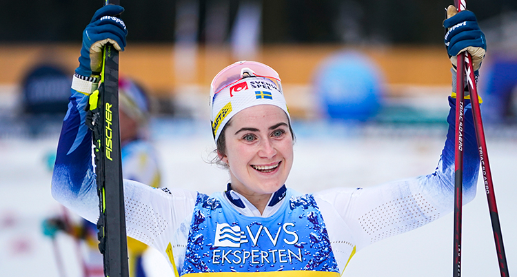 Hon är glad och sträcker upp armarna som hon håller skidor och stavar med.