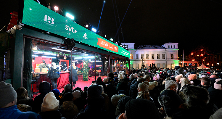 En bur av glas med gröna skyltar står på torget. Många människor står framför och tittar.