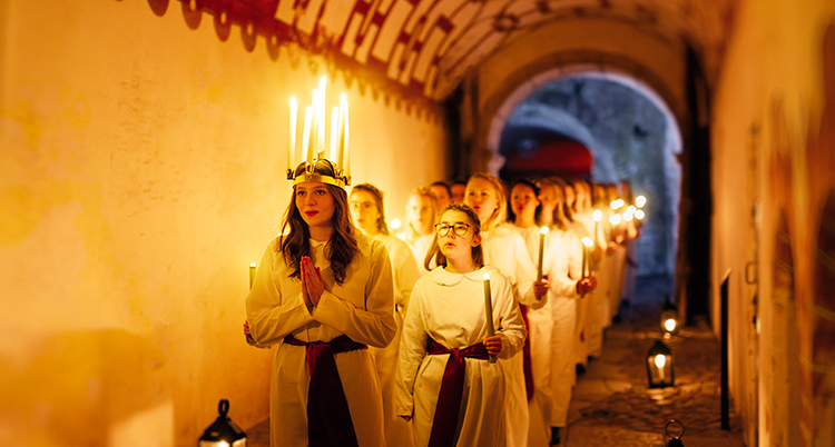 Ett luciatåg går genom en gång inomhus. Lucia går först och har en krona med ljus. Tärnor går efter med vita klänningar och ljus i händerna.
