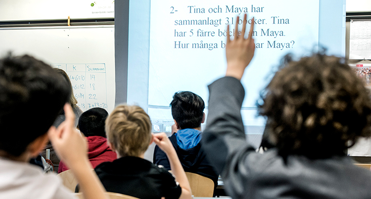 Barn som sitter i ett klassrum. Ett barn räcker upp en hand.