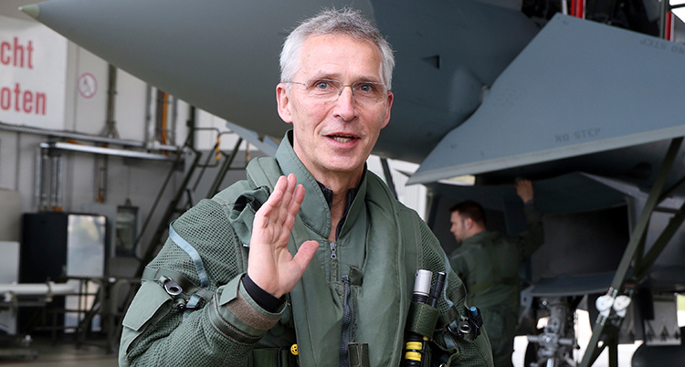 Han står framför ett grått militärt flygplan. Han tittar in i kameran och vinkar med sin höra hand.