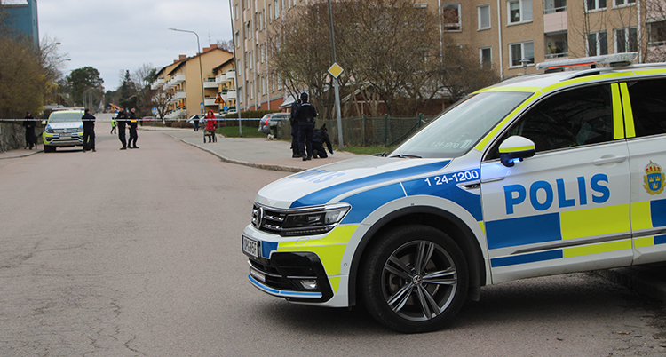 En polisbil på en gata. I bakgrunden står flera poliser vid ett plastband.