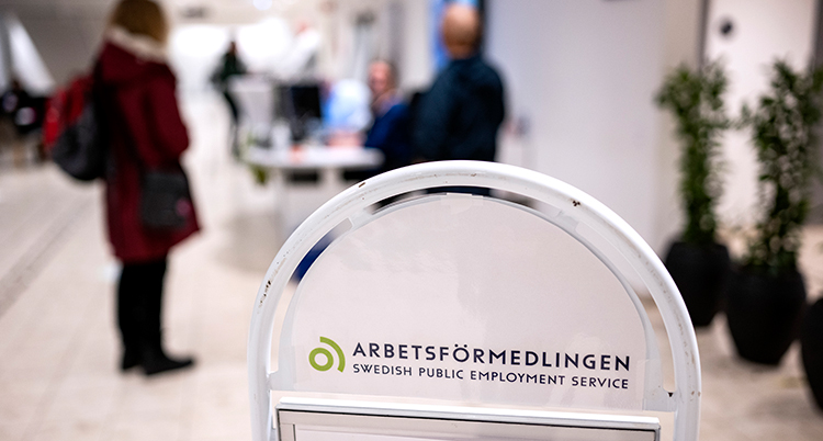 En skylt från Arbetsförmedlingen. I bakgrunden syns människor som besöker Arbetsförmedlingen.