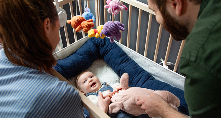 De lutar sig ner över en spjälsäng. Där ligger en baby. En leksak hänger över sängen.
