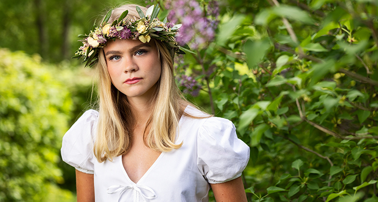 Hon har en krans av blommor på huvudet. Hon ser allvarlig ut.
