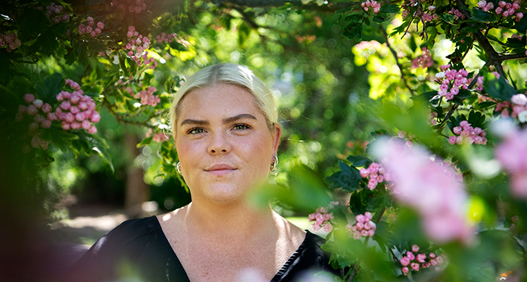 Hon har blont hår . Hon ler. Hon står bland gröna blad och rosa blomor.