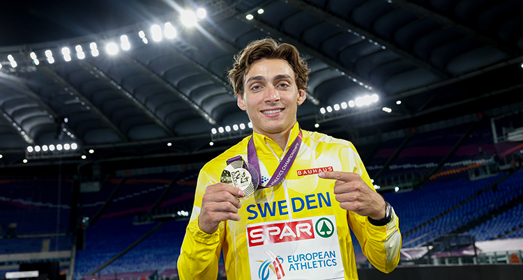 Han står mitt i en stor arena med lampor. Han håller upp sin medalj och pekar på den.