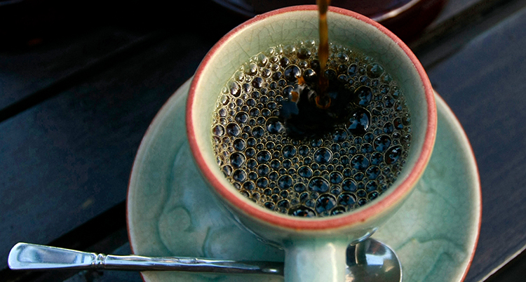 Någon häller kaffe i en kaffekopp med en sked vid sidan om.