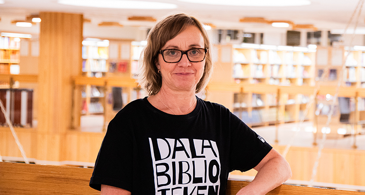 Hon står i ett bibliotek. Hon tittar in i kameran.