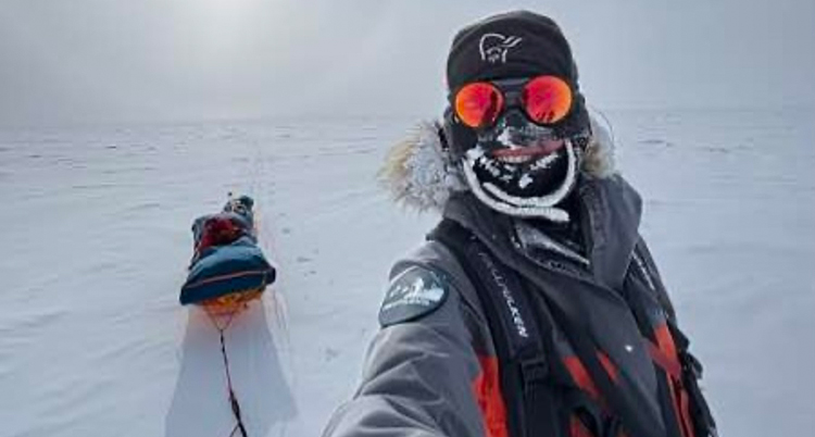 Hon har tjocka kläder och en huva och glasögon. Det är vitt av snö.. Bakom syns en pulka.