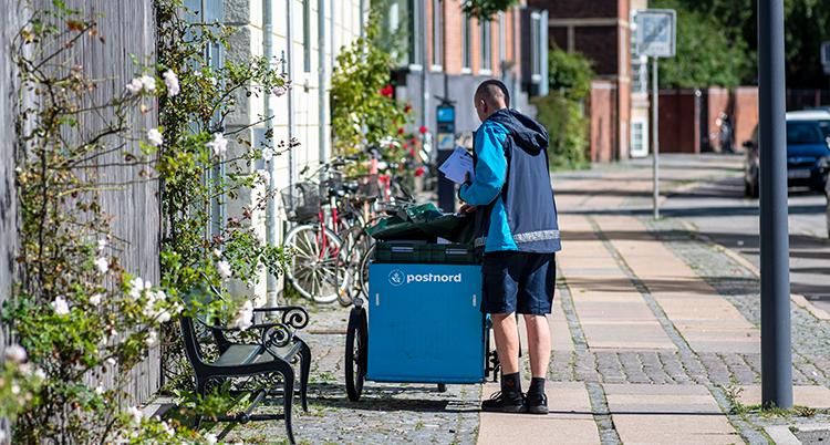 En person står på en trottoar i en gata, bredvid en vagn med brev.