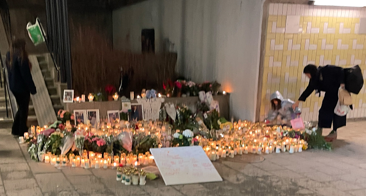 En plats fylld med blommor och tända ljus. Några personer står vid platsen.