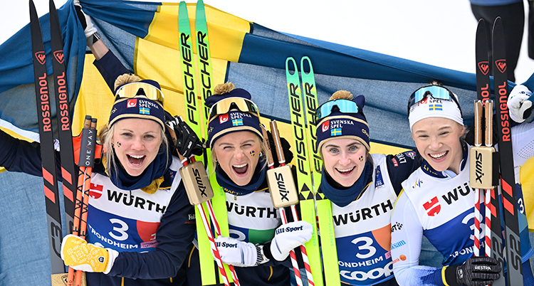 De står bredvid varandra och håller i sina skidor och stavar. Bakom syns en svensk flagga.