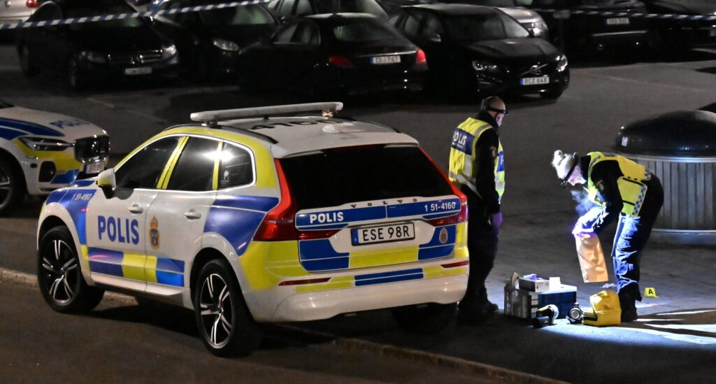 Poliser vid en polisbil jobbar på en parkeringsplats. Det är kväll och de lyser med lampor.