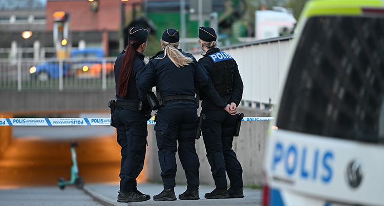 Tre poliser står med ryggen mot kameran. En polisbil syns till höger.