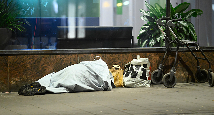 En person ligger på trottoaren. Personens rullator och väska står bredvid.