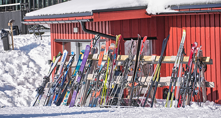 Många skidor står i ett ställ utanför ett rött hus. Det är snö omkring.