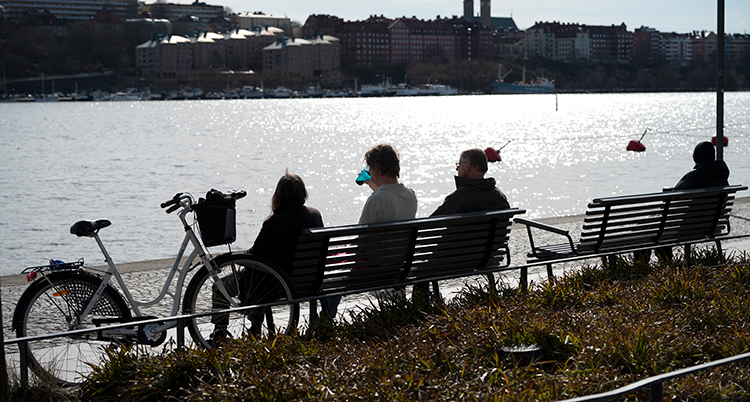 Folk sitter på en bänk i solen. De ser på vattnet.