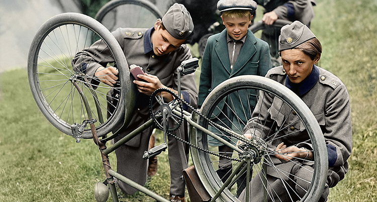 Två soldater i gråa uniformer lagar en cykel som är upp och ned. En liten kille i keps och kavaj tittar på.