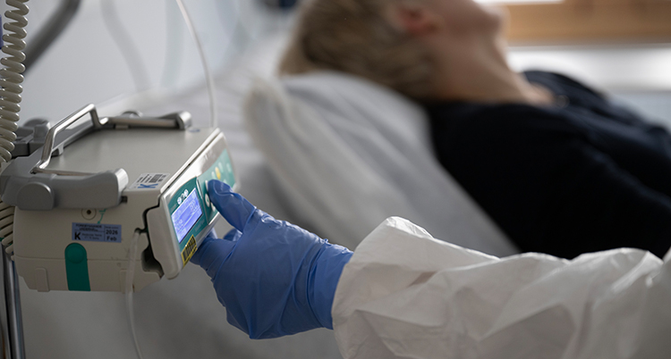En person ligger i en säng på ett sjukhus. En sjuksköterska trycker på en knapp till patients dropp.