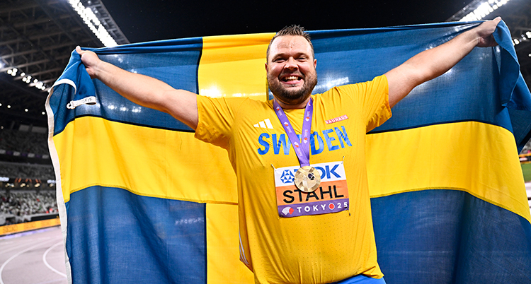 Han håller i en svensk flagga. Han ler. Han har en medalj om halsen.