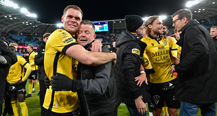 Efter en match i fotboll. Flera personer är på planen. En spelare kramar en tränare.