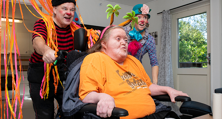 Två clowner skjutsar en kvinna som sitter i en rullstol.
