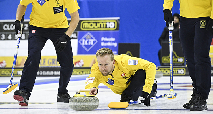 Han glider utsträckt på isen med en curlingsten i handen. Andra spelare är vid sidan om.