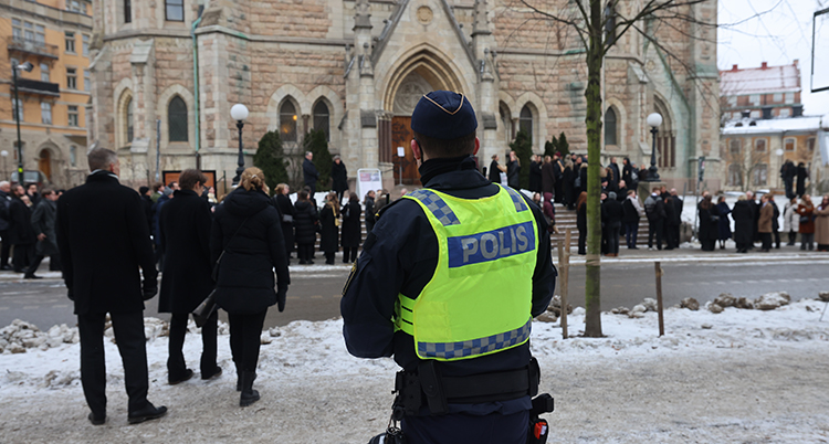 Ryggen av en polis med reflexväst. Många människor går mot kyrkans port.