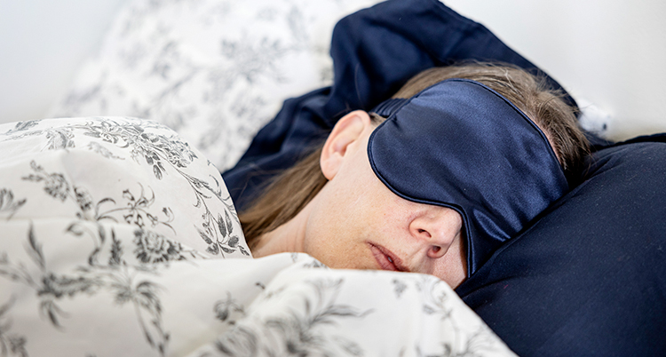 En kvinna ligger i en säng. Hon har en sovmask över ögonen.