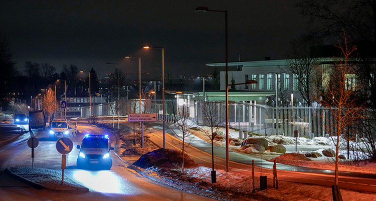 Det är mörkt ute. En stor byggnad syns. DEt lyser i fönstren. Det är staket omkring. Polisbilar står utanför staketet.