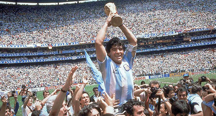 På en fotbollsplan. Människor lyfter upp Maradona. Han håller VM-trofén i sina händer.