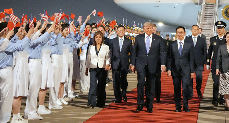 På en flygplats. Donald Trump går på en röd matta från ett flygplan. Han går med flera andra. Bredvid står personer och viftar med flaggor.