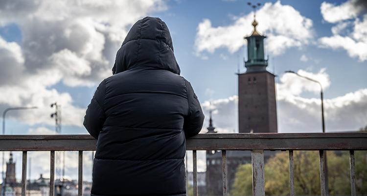 En person står vid en bro. Personen har en stor jacka och ryggen mot kameran.