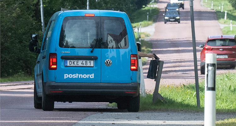 En bil står på en väg intill brevlådor. En hand sträcks ut och lägger ner post i lådan.