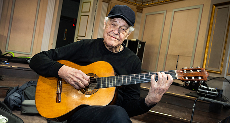 Han sitter ner och spelar gitarr.