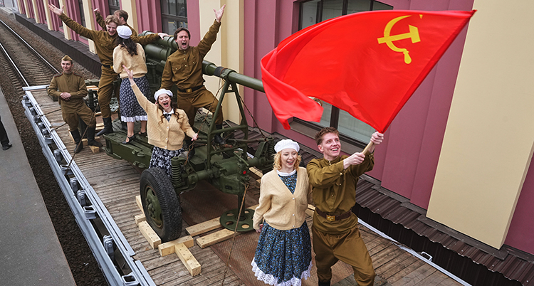 Några soldater och kvinnor står på en vagn. De viftar med en sovjetisk flagga och är glada.