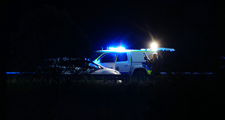 Det är mörkt ute. En vit bil står på en väg. Bredvid står en polisbil. Några poliser är också där.