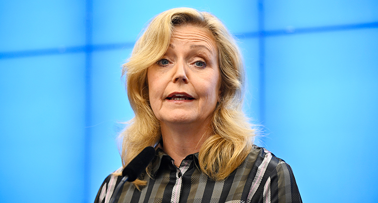 Hon står och pratar på en träff med journalister. Hon har ljust långt hår och en rutig skjorta.