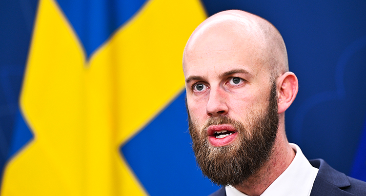 Han pratar på ett möte med journalister. Bakom honom syns en svensk flagga.