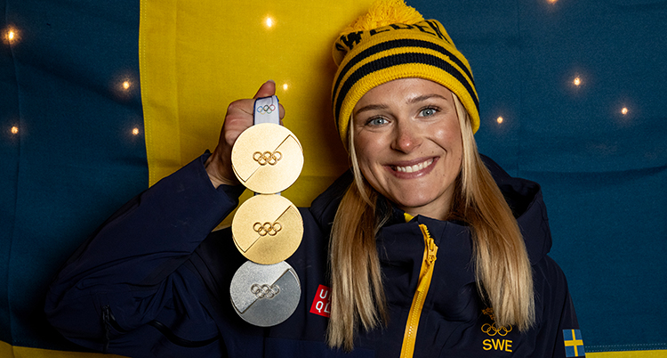 En nära bild. Hon har en mössa på sig. Hon håller upp tre medaljer.