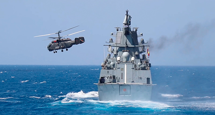 Ett grått fartyg för militärer som är ute på havet. En helikopter flyger bredvid.