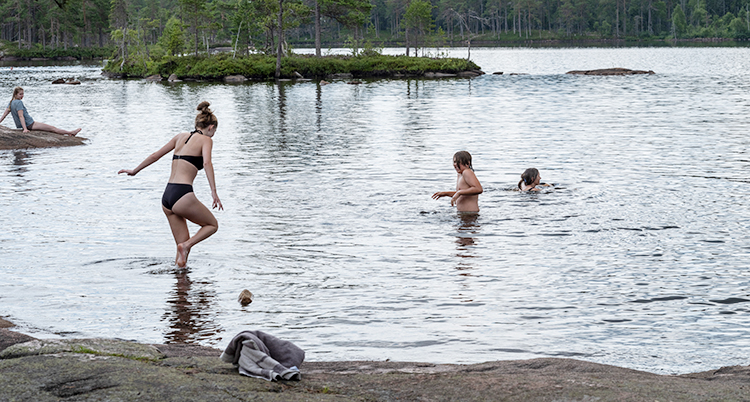 Två personer är i vattnet. En tredje är på väg ner. En fjärde sitter på en klippa en bit bort.