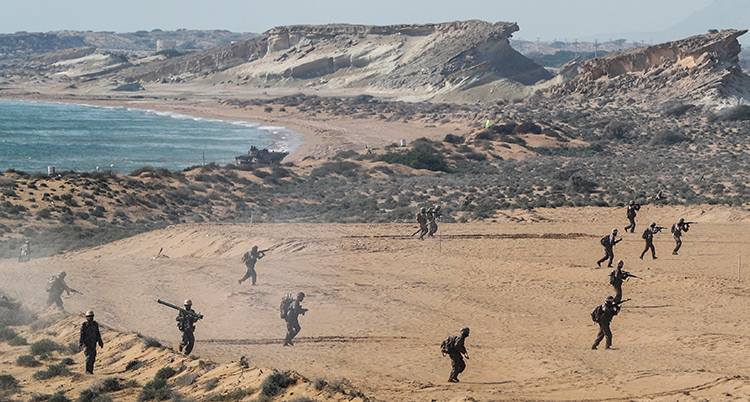 Militärer springer i sand. I bakgrunden syns hav och berg.